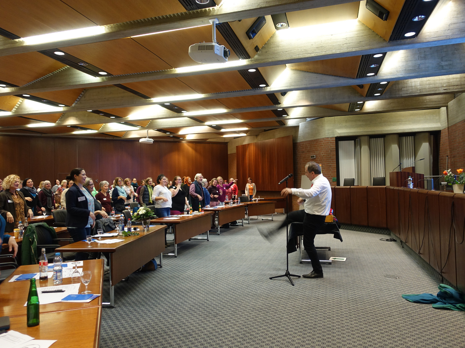 Eine Menschenmenge steht in einem Konferenzraum, während ein Redner am Mikrofon spricht und energetisch eine Darbietung zeigt. Tische sind mit Notizen und Getränken gedeckt. Die Atmosphäre ist lebhaft und engagiert.