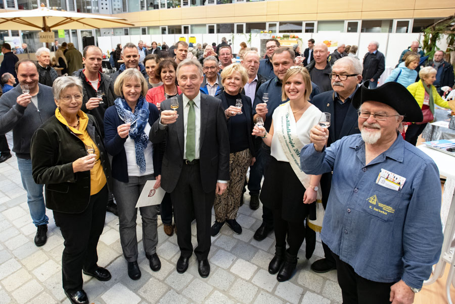 Eine Gruppe von Menschen steht zusammen und hebt Gläser zum Anstoßen. Sie lächeln und scheinen eine Feier oder ein besonderes Ereignis zu genießen. Im Hintergrund sind weitere Gäste und eine helle, freundliche Umgebung zu sehen.
