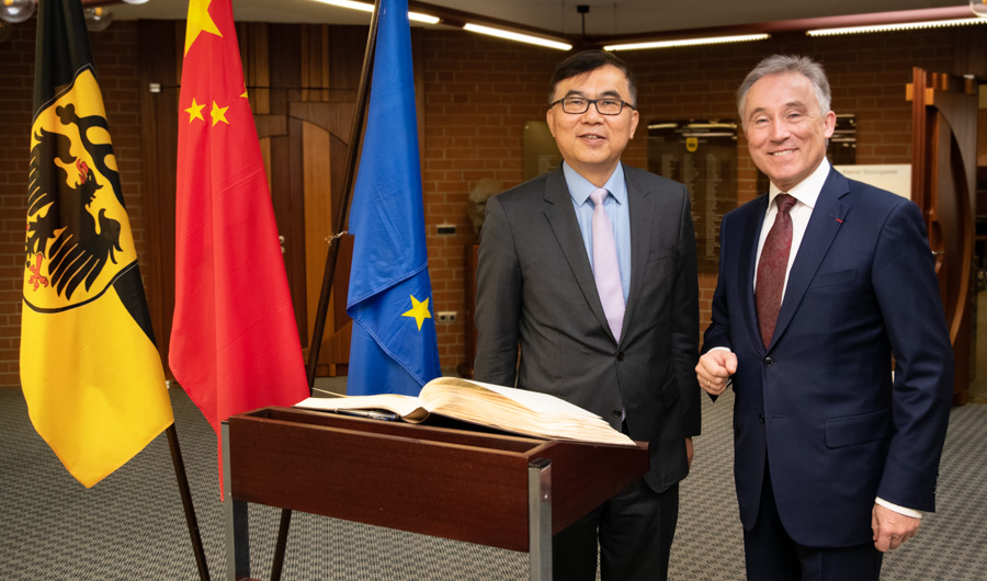 Zwei Männer stehen in einem Raum, umgeben von Flaggen Deutschlands, Chinas und der Europäischen Union. Sie lächeln und präsentieren ein aufgeschlagenes Buch, das auf einem Tisch liegt. Die Szene vermittelt ein Gefühl der Zusammenarbeit und des Austauschs zwischen den Nationen.