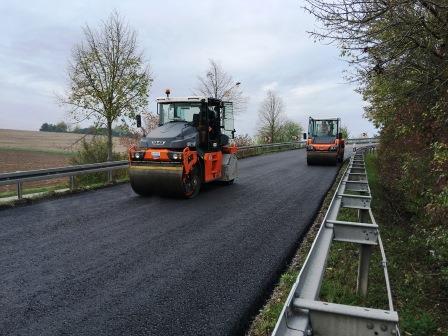 Zwei Straßenwalzen arbeiten an der Asphaltierung einer Straße. Im Hintergrund sind Bäume und eine offene Feldlandschaft zu sehen. Der Asphalt ist frisch und noch schwarz, während die Maschinen sich entlang der Straßenkante bewegen.