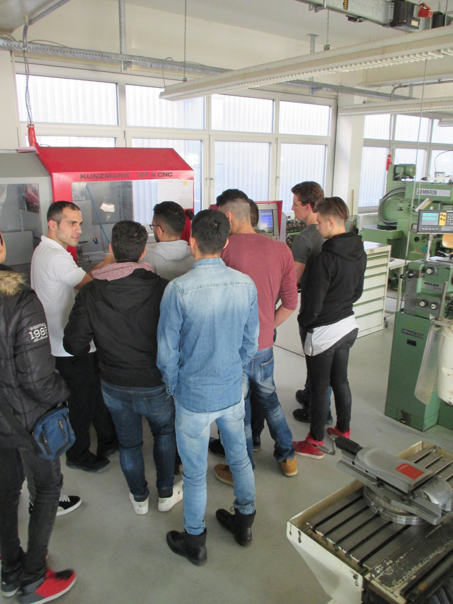 A diverse group of individuals attentively listens to an instructor in a workshop setting. The instructor stands in front of a machine, explaining its operation, while students are gathered closely around, engaged in the discussion. Industrial equipment is visible in the background.