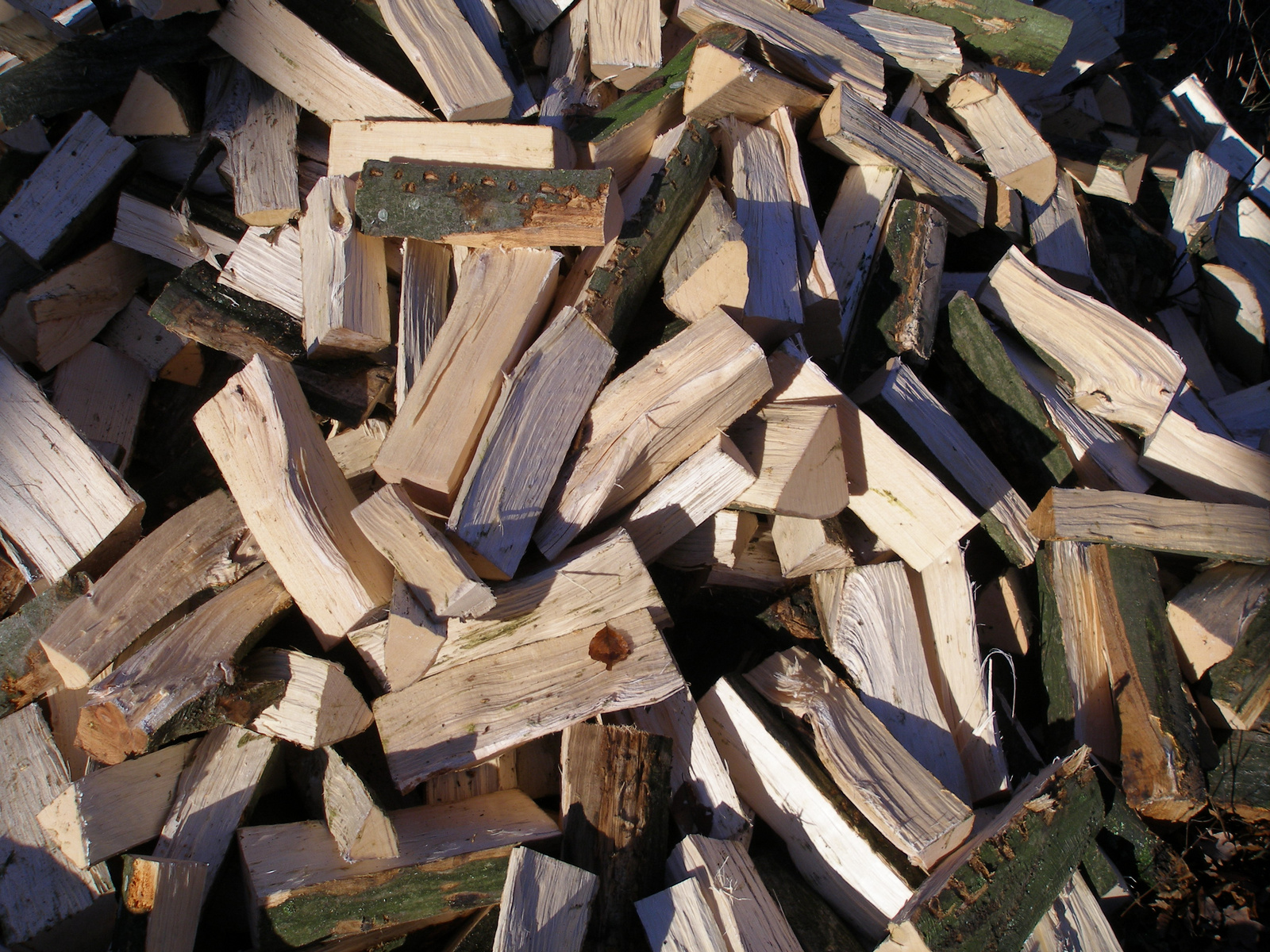 A close-up view of a pile of firewood logs in various sizes, predominantly light-colored with some pieces showing dark green bark. The stacked logs are randomly arranged, creating a textured and layered appearance.