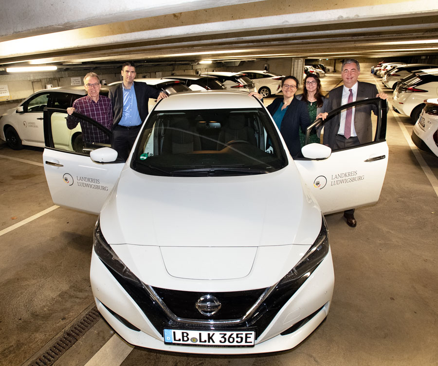 Eine Gruppe von fünf Personen steht um ein weißes Elektroauto in einer Tiefgarage. Zwei Personen halten die Tür des Fahrzeugs offen. Das Auto gehört zum Landkreis Ludwigsburg. Hintergrund mit anderen parkenden Fahrzeugen, die den Fokus auf die Gruppe und das Auto legen.
