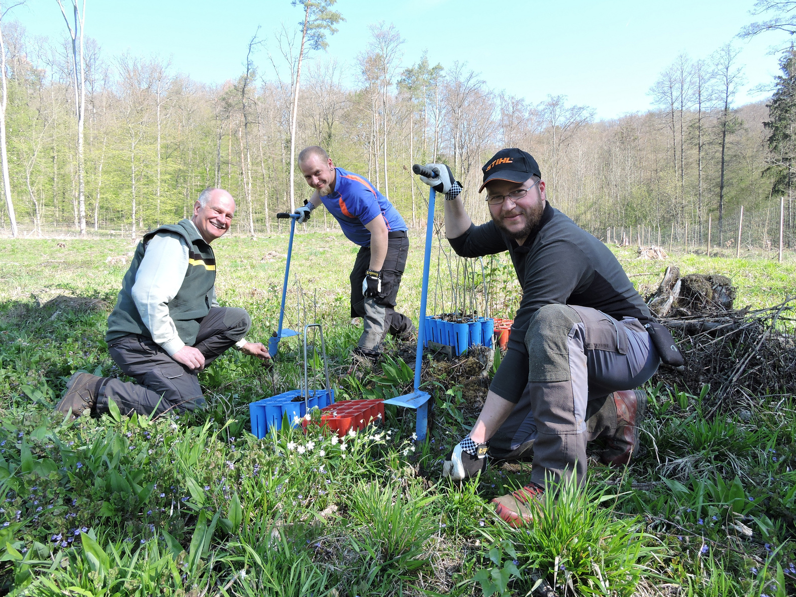 Drei Personen knien auf einer Wiese und pflanzen junge Bäume. Sie tragen Arbeitskleidung und lachen, während sie ihre Werkzeuge nutzen. Hinter ihnen ist ein Wald mit frischem Grün sichtbar. Der Himmel ist klar und die Szene vermittelt einen Eindruck von Gemeinschaft und Naturschutz.