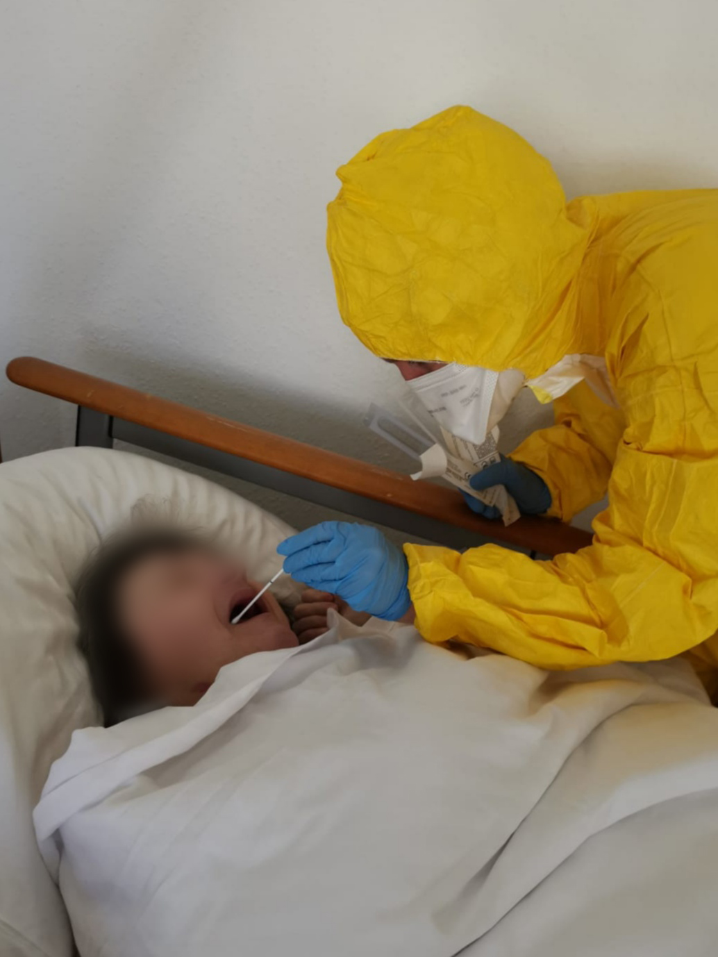 Eine Person in einem gelben Schutzanzug führt einen Test mit einem Abstrich bei einem im Bett liegenden Patienten durch, der in eine Decke gehüllt ist. Der Patient ist nicht klar erkennbar. Die Szene vermittelt einen medizinischen Kontext, möglicherweise im Zusammenhang mit einer Gesundheitsuntersuchung.