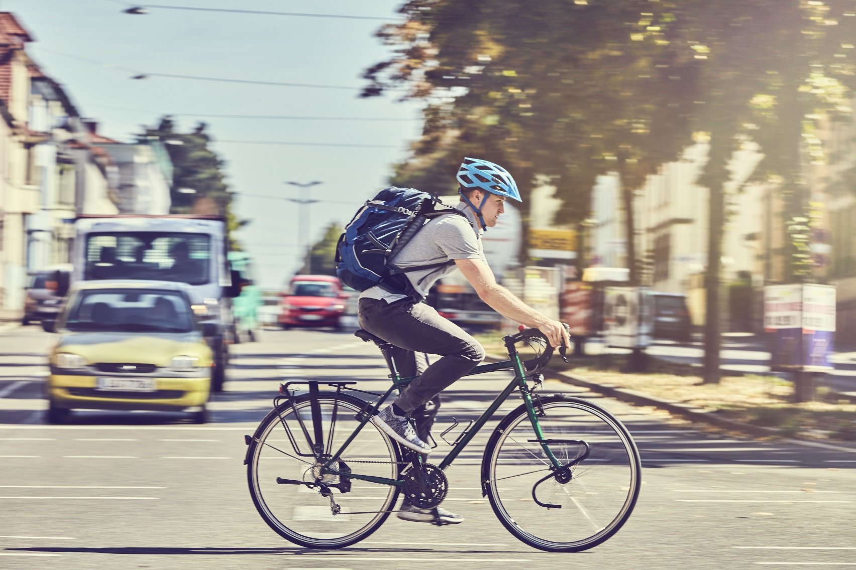 Ein Mann fährt mit einem Fahrrad auf einer Stadtstraße. Er trägt einen blauen Helm und einen Rucksack. Im Hintergrund sind Autos und Bäume zu sehen, die eine urbanen Umgebung widerspiegeln.
