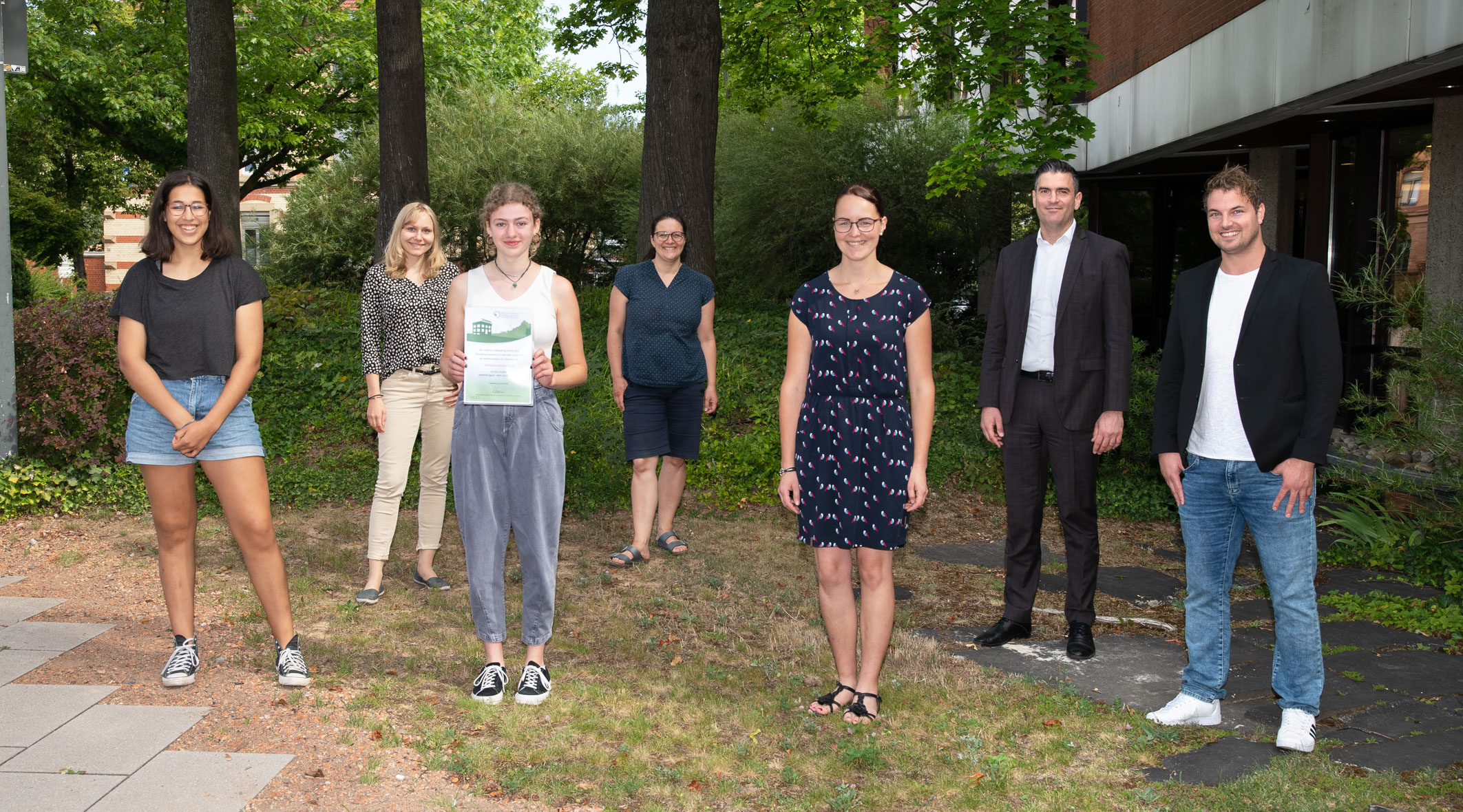 Die Abbildung zeigt sieben Personen, die im Freien auf einer Wiese stehen. Einige halten Dokumente in den Händen. Der Hintergrund ist grün mit Bäumen und Büschen, während der Boden mit Kies und Rasen bedeckt ist. Es sieht nach einer informellen Veranstaltung aus.