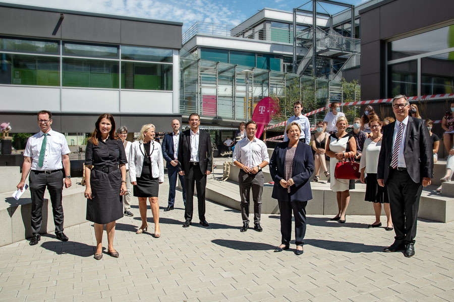 Gruppenszene von Personen, die im Freien stehen und in die Kamera lächeln. Im Hintergrund sind moderne Gebäude und eine Menschenmenge zu sehen. Die Personen tragen formelle Kleidung und scheinen an einer Veranstaltung teilzunehmen.