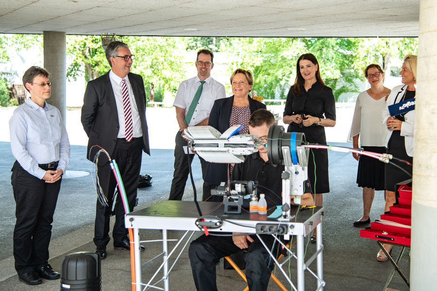 Eine Gruppe von Personen beobachtet eine technische Demonstration. Im Vordergrund sitzt eine Person an einem Tisch mit einem Gerät, während die anderen Teilnehmer, darunter einige in formeller Kleidung, aufmerksam zuschauen. Die Umgebung ist hell und offen, mit Bäumen im Hintergrund.