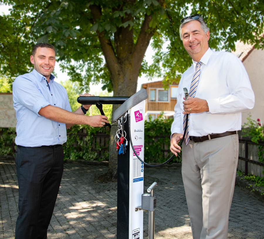 Zwei Männer stehen an einer Säule mit einem Schlüsselanhänger. Der Mann links trägt ein hellblaues Hemd, der rechts ein weißes Hemd mit Krawatte. Im Hintergrund ist ein Baum sowie ein österlicher Platz zu sehen. Beide wirken freundlich und lächeln.