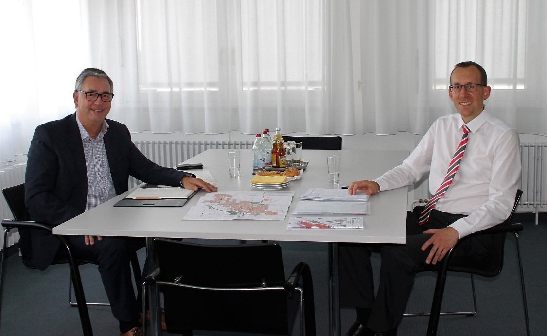 Zwei Männer sitzen an einem Tisch in einem Büro. Der Mann links trägt einen Anzug und eine Brille, der Mann rechts hat ein Hemd mit einer gestreiften Krawatte. Auf dem Tisch liegen Unterlagen und Wassergläser. Im Hintergrund sind große Fenster mit Vorhängen.