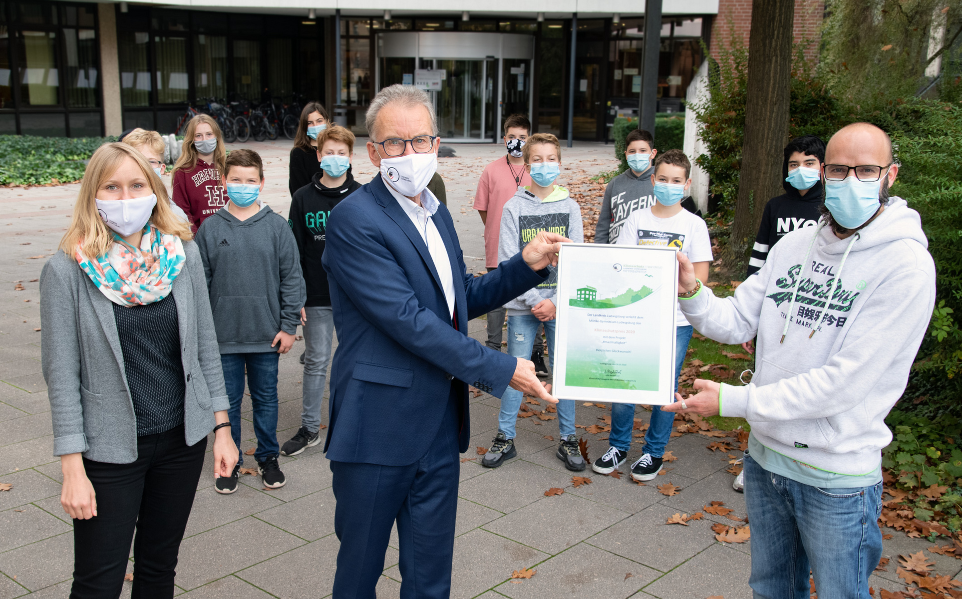 Eine Gruppe von Schülern und Lehrern steht im Freien, alle tragen Mund-Nasen-Schutz. Ein Lehrer überreicht eine Urkunde an einen Schüler. Im Hintergrund ist ein Schulgebäude zu sehen. Die Stimmung wirkt feierlich und gemeinschaftlich.