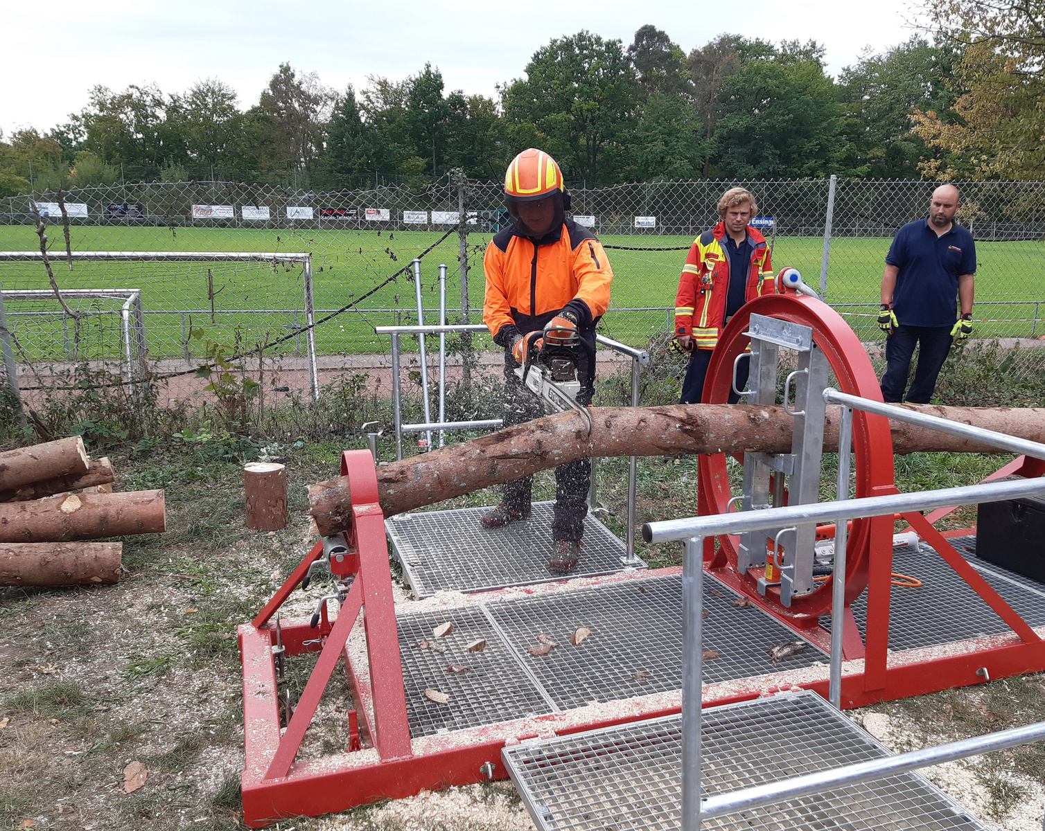 Ein Mann in orangefarbener Sicherheitskleidung und Helm bedient eine motorsägengestützte Maschine, um einen dicken Baumstamm zu schneiden. Im Hintergrund stehen zwei weitere Personen und eine große, grüne Rasenfläche ist sichtbar. Es handelt sich um eine Demonstration für die Holzverarbeitung.