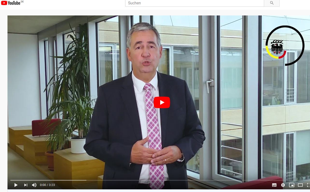 A man in a suit stands in an office space with large windows, speaking directly to the camera. He gestures with his hands as he presents information, while a red YouTube play button appears in the center of the image, indicating a video format.