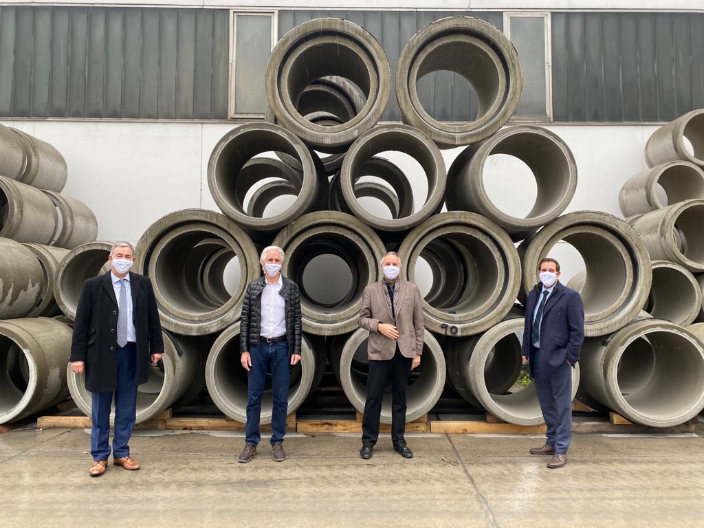 Vier Personen stehen vor einem Stapel aus großen Betonrohren. Alle tragen Masken. Die Gruppe scheint ein wichtiges Treffen oder eine Besichtigung in einem industriellen Umfeld zu haben.