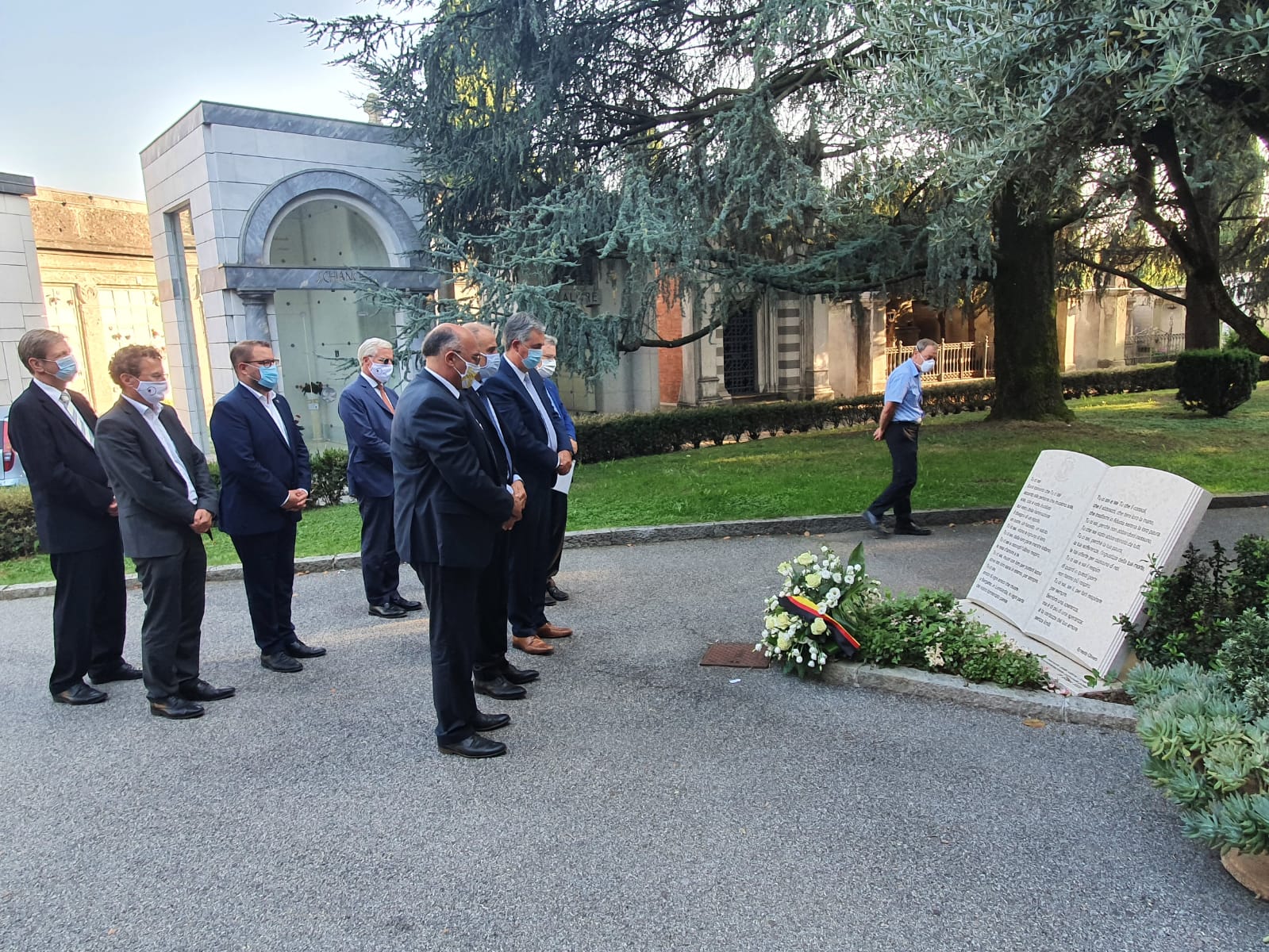 Eine Gruppe von sieben Menschen in Anzügen und mit Mundschutz steht in einem Friedhofsbereich. Sie schauen auf eine Gedenktafel und eine Kranzniederlegung. Im Hintergrund sind Bäume und mausoleumsartige Strukturen zu sehen. Die Szene vermittelt Respekt und Gedenken.