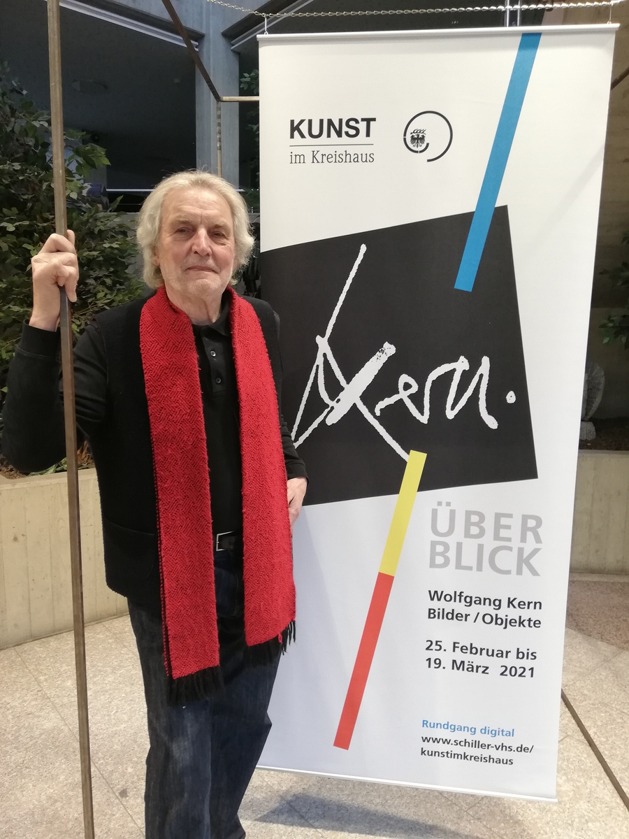 Ein älterer Mann mit langen grauen Haaren und einem roten Schal steht neben einem Banner. Das Banner zeigt den Schriftzug „Kern“ und Informationen zur Ausstellung von Wolfgang Kern mit dem Titel „Überblick“, die vom 25. Februar bis 19. März 2021 im Kreishaus stattfindet.