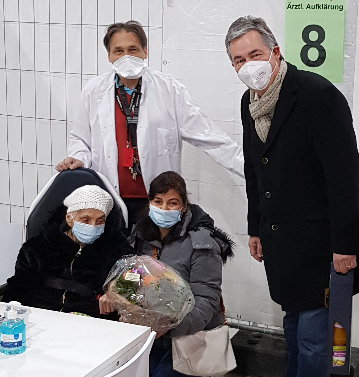 Eine ältere Frau sitzt in einem Stuhl mit einer Blume in der Hand. Neben ihr steht eine weitere Frau, beide tragen Mund-Nasen-Schutz. Im Hintergrund sind zwei Männer, einer im weißen Kittel und der andere in einem dunklen Mantel, ebenfalls mit Maske. An der Wand hängt ein Schild mit der Nummer 8.