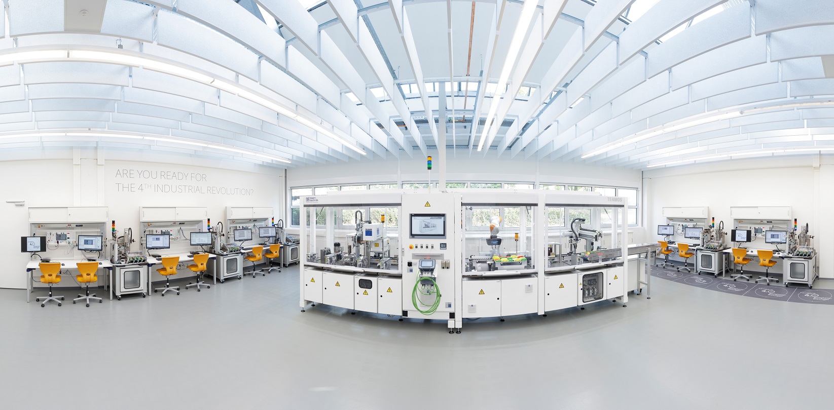 Das Bild zeigt einen modernen, hellen Raum mit mehreren Arbeitsstationen und Maschinen. In der Mitte befindet sich eine große Einheit mit digitalen Displays. An den Seiten stehen orangefarbene Arbeitsplätze, die für industrielle Anwendungen eingerichtet sind. Die Atmosphäre vermittelt Fortschritt und Technologie.