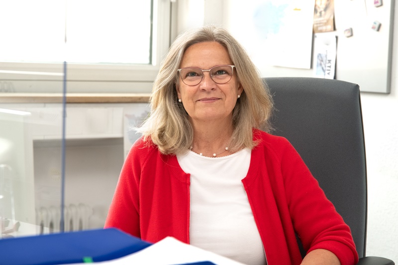 Eine lächelnde Frau mit blonden, lockigen Haaren und Brille sitzt an einem Schreibtisch. Sie trägt einen roten Pullover über einem weißen Oberteil. Im Hintergrund sind einige Büroartikel und eine Fensteransicht zu sehen.