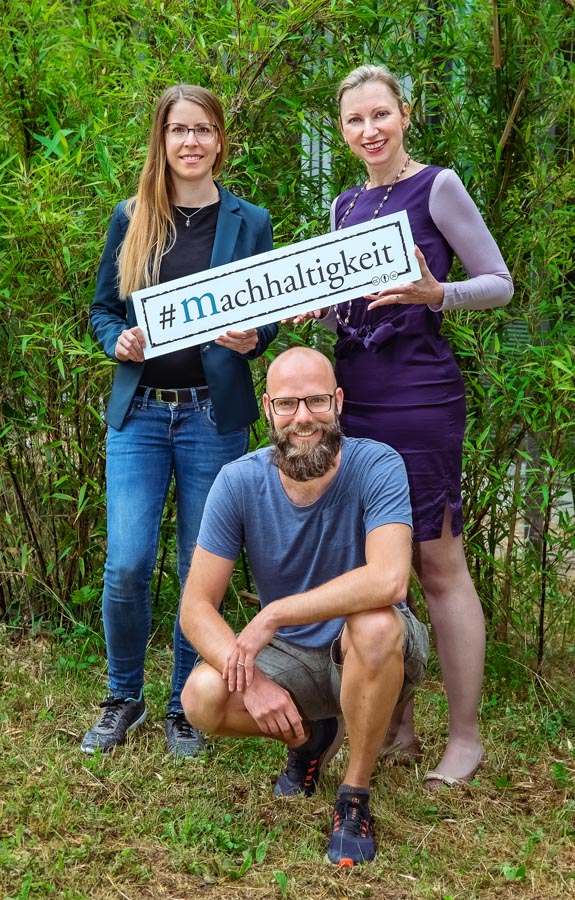 Drei Personen posieren vor einem grünen Hintergrund. Eine Frau hält ein Schild mit dem Hashtag "#Nachhaltigkeit". Die Szene vermittelt das Thema Nachhaltigkeit und Umweltbewusstsein.