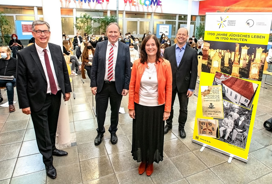 Quatre personnes se tiennent debout dans un espace public, souriant et vêtues de costumes formels. À droite, une affiche colorée présente un projet sur la vie juive, intitulé « 1700 Jahre jüdisches Leben in 1700 Minuten ». En arrière-plan, un groupe de personnes assises regarde.
