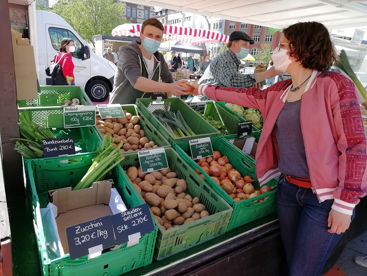 Ein Verkäufer reicht einer Kundin eine Tüte mit frischem Gemüse. Im Vordergrund sind Kisten mit Zucchini, Kartoffeln und Äpfeln sichtbar. Beide tragen Masken. Der Markt hat ein lebhaftes Ambiente mit weiteren Ständen im Hintergrund.