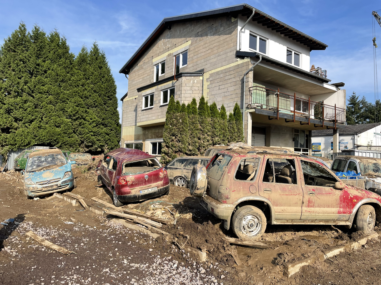 Ein beschädigtes Wohnhaus steht neben einer Reihe von verschmutzten Autos, die in schlammigem Gelände stehen. Die Fahrzeuge sind mit Schmutz und Trümmern bedeckt. Im Hintergrund sind grüne Bäume zu sehen, die die Szene umrahmen.