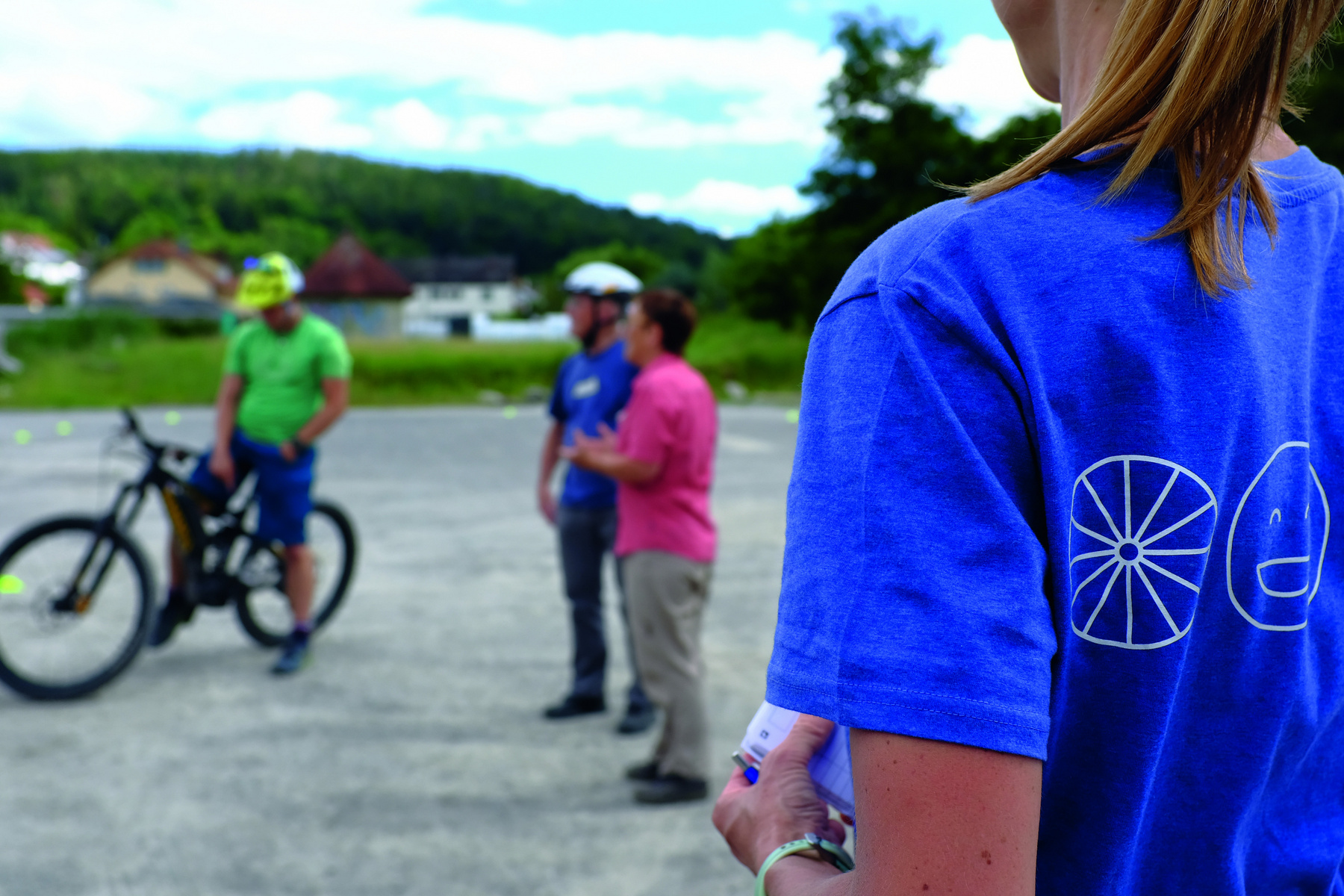 Auf dem Bild sieht man eine Gruppe von Personen, die sich im Freien versammeln. Eine Person trägt ein blaues T-Shirt mit einem grafischen Motiv. Im Hintergrund stehen zwei Männer, einer auf einem Fahrrad, während sie mit einer Frau sprechen, die Anweisungen gibt.