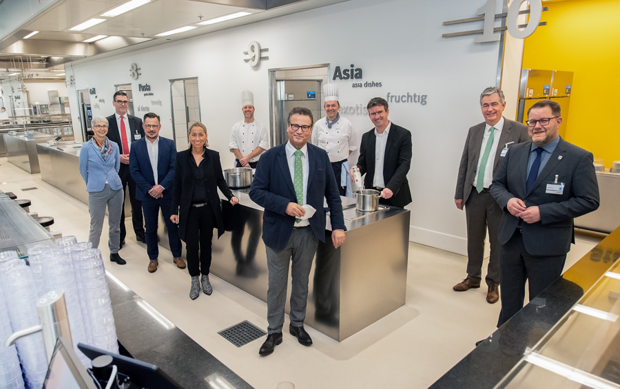 Eine Gruppe von zwölf Personen steht in einer modernen Küche, umgeben von Kochbereichen. Einige Personen tragen Kochjacken. Die Atmosphäre wirkt professionell und freundlich, als ob ein Event oder eine Eröffnung stattfindet. Im Hintergrund sind Beschriftungen zu sehen, die verschiedene Küchenstile angeben.