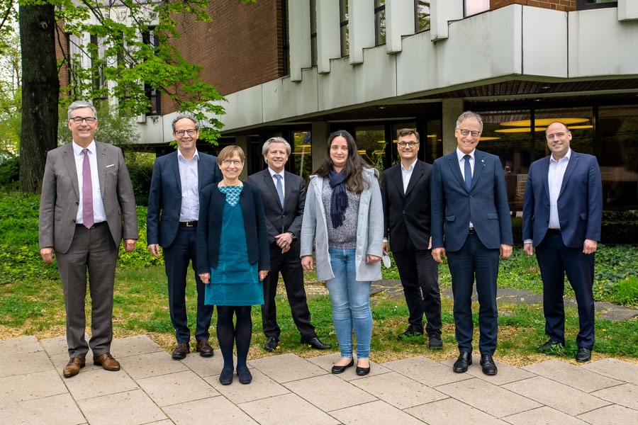 Eine Gruppe von acht Personen steht draußen in einer Gartenlandschaft. Sie sind in formeller Kleidung gekleidet und lächeln in die Kamera. Die Szene vermittelt ein Gefühl von Zusammengehörigkeit und Professionalität.