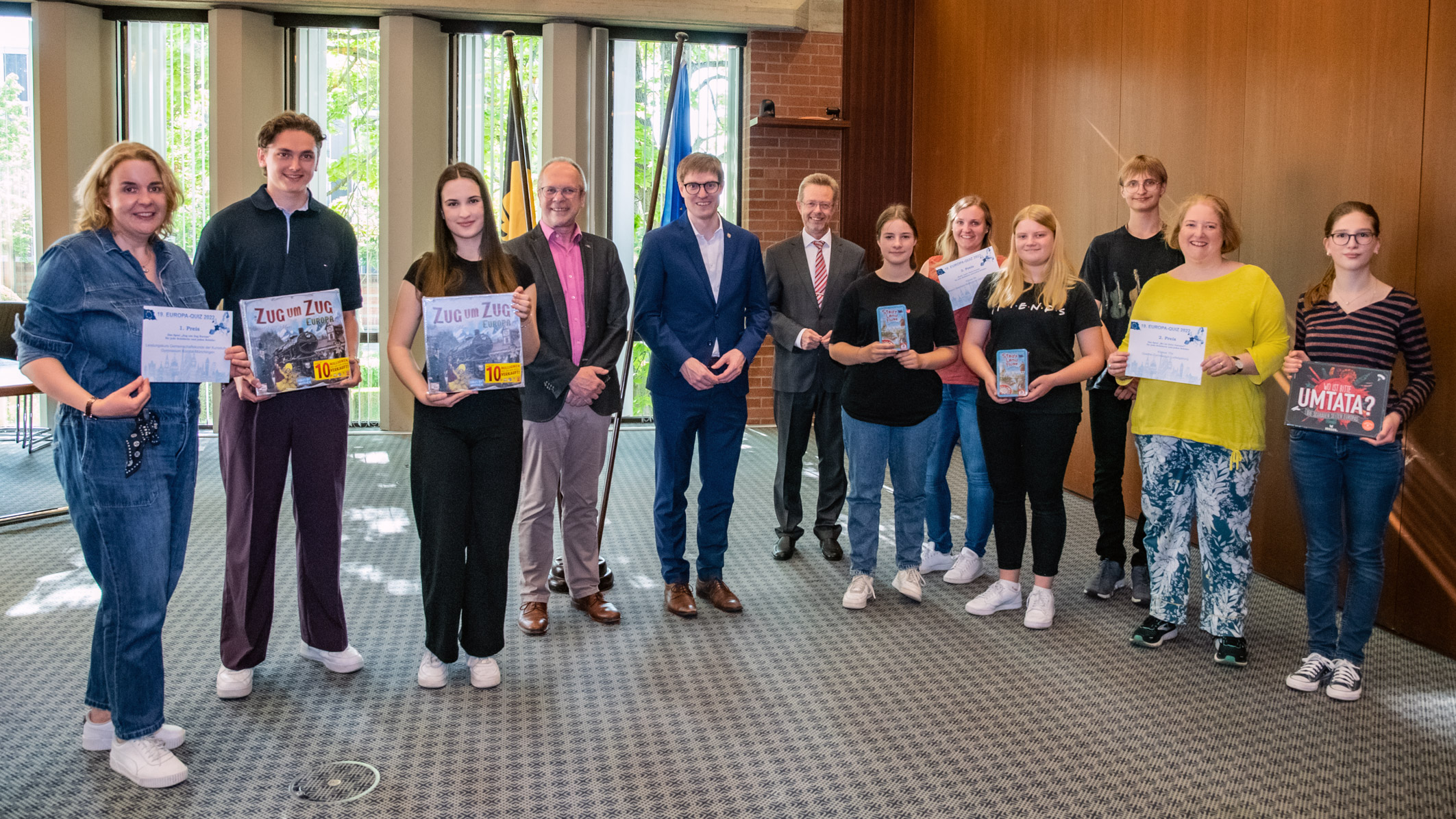 Gruppenvorstellung in einem Raum mit großen Fenstern. Eine Gruppe von zehn Personen steht in zwei Reihen. Einige halten Preisauszeichnungen oder Bücher in der Hand. Im Hintergrund stehen weitere Menschen und lächeln. Die Atmosphäre wirkt festlich und erfreulich.