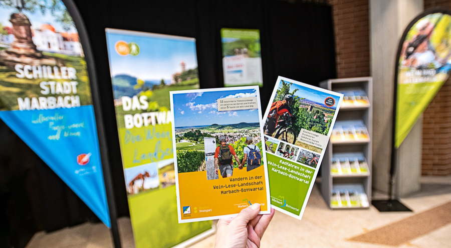 Eine Hand hält zwei Broschüren über Wanderangebote in einer Weinbau-Landschaft. Im Hintergrund befinden sich farbige Plakate mit Informationen über die Region Marbach und touristische Aktivitäten. Die Atmosphäre vermittelt ein einladendes Gefühl für Natur- und Wanderfreunde.