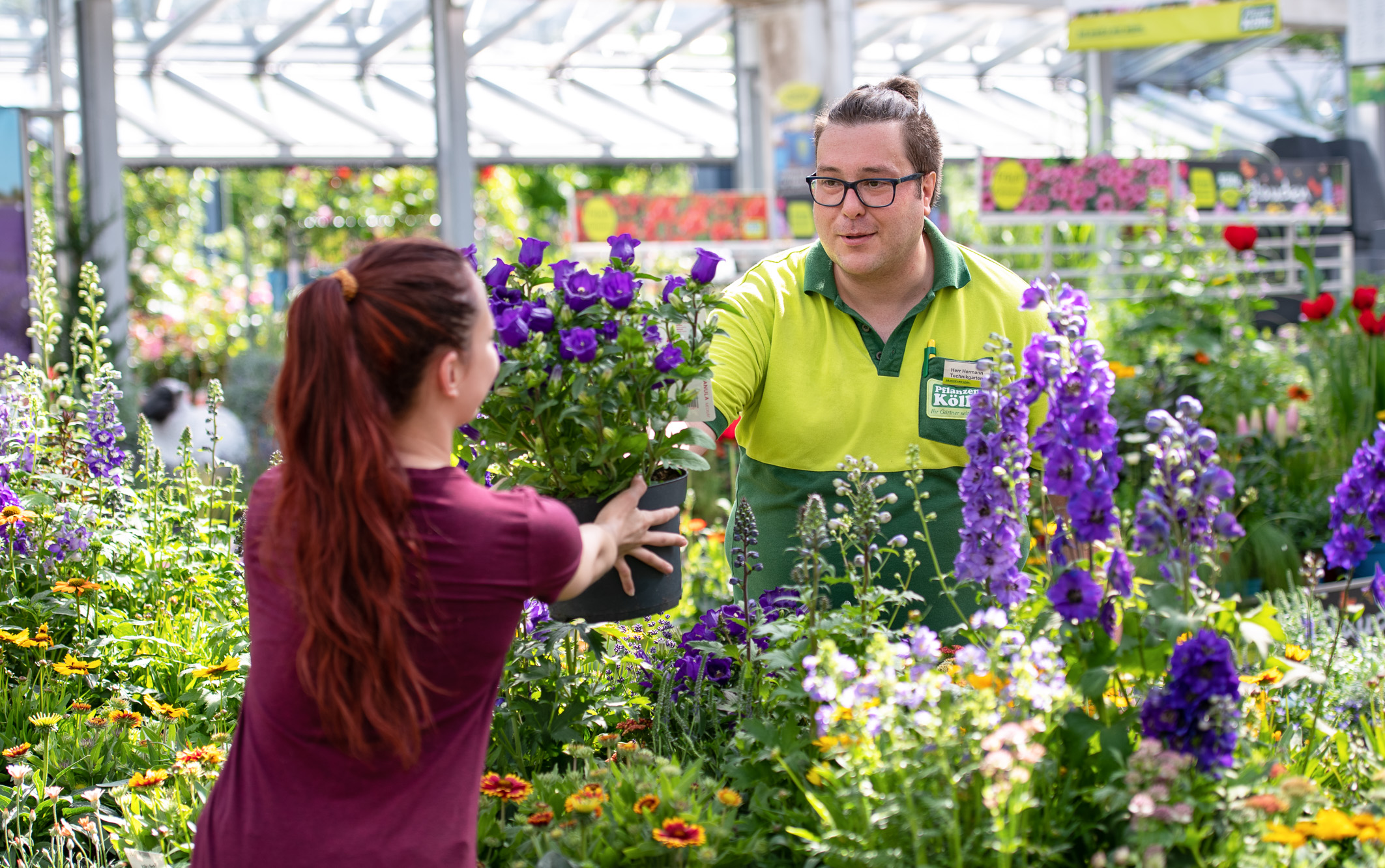 Ein Mitarbeiter eines Gartencenters übergibt einer Kundin einen Topf mit lila Blumen. Hinter ihnen sind viele bunte Pflanzen und Blumen zu sehen. Das Bild vermittelt eine freundliche Atmosphäre des Floristik- und Gartenhandels.
