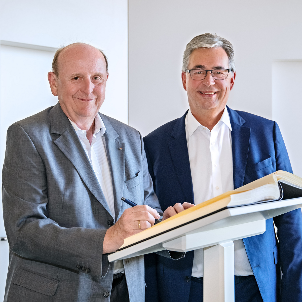 Zwei Männer stehen nebeneinander, lächelnd in einem hellen Raum. Der Mann links trägt einen grauen Anzug und hält einen Stift, während der Mann rechts ein weißes Hemd und einen blauen Blazer trägt. Auf einem Tisch zwischen ihnen liegt ein aufgeschlagenes Buch.
