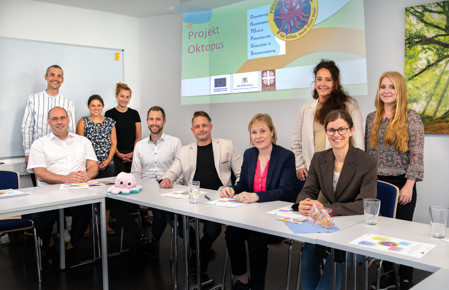 Eine Gruppe von zehn Personen sitzt und steht um einen Tisch. Im Hintergrund ist eine Präsentation mit dem Titel „Projekt Oktopus“ zu sehen. Die Teilnehmer scheinen an einem Arbeitsmeeting oder einer Präsentation teilzunehmen. Auf dem Tisch liegen verschiedene Unterlagen und ein Plüschtier.