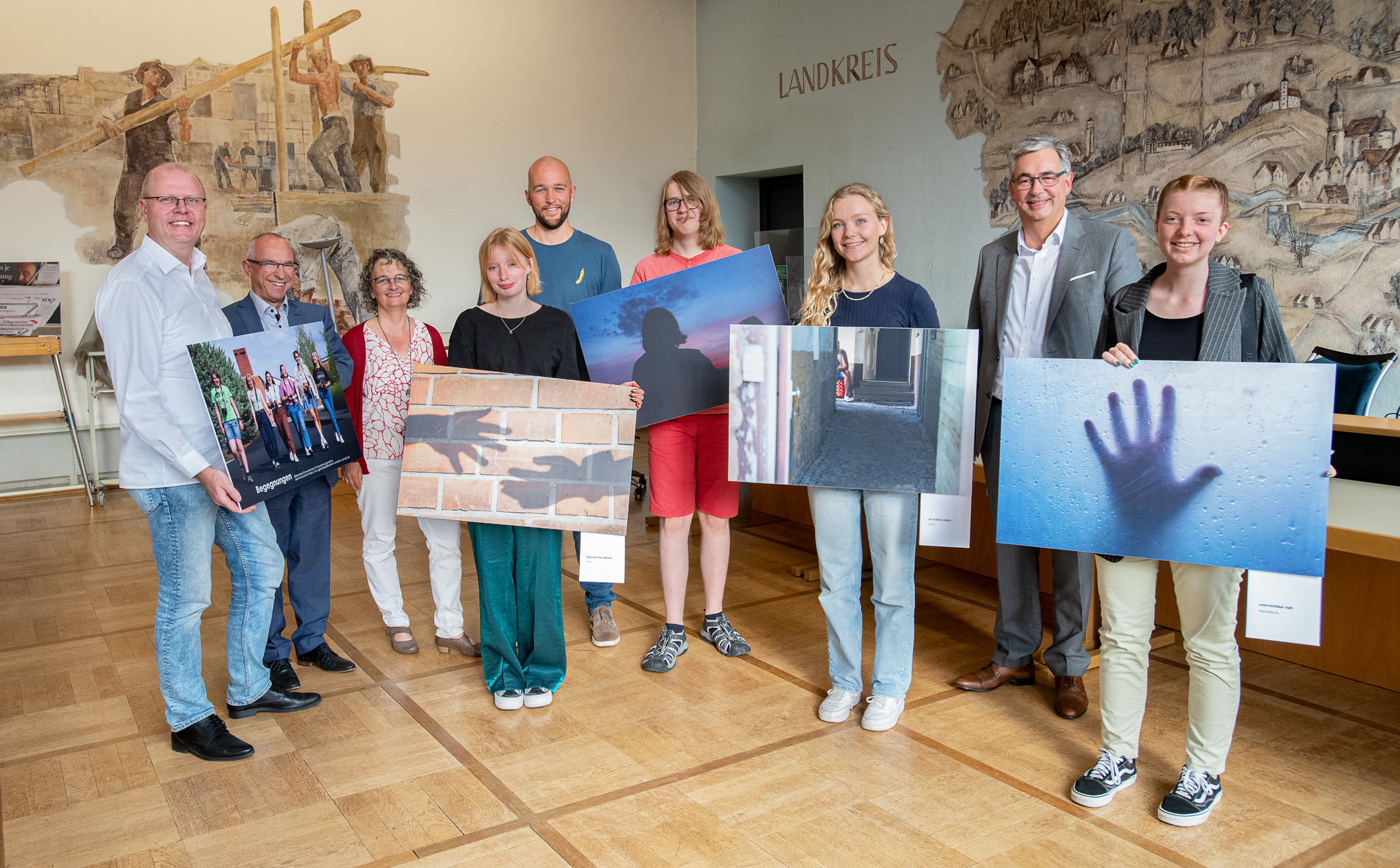 Eine Gruppe von sieben Personen steht in einem Raum und hält verschiedene große Fotos in den Händen. Die Bilder zeigen unterschiedliche Motive, während im Hintergrund eine Wand mit einem historischen Wandgemälde zu sehen ist.