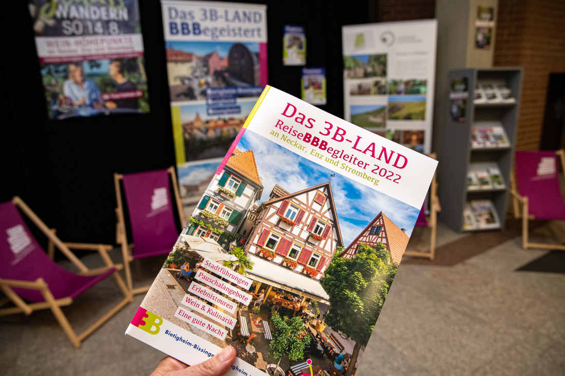 Ein Reiseführer für das 3B-Land, inklusive Bilder von malerischen Gebäuden. Der Titel „ReiseBBegleiter 2022“ ist prominent auf dem Cover abgebildet. Im Hintergrund sind Informationsstände mit weiteren Materialien und Plakaten zur Region sichtbar.