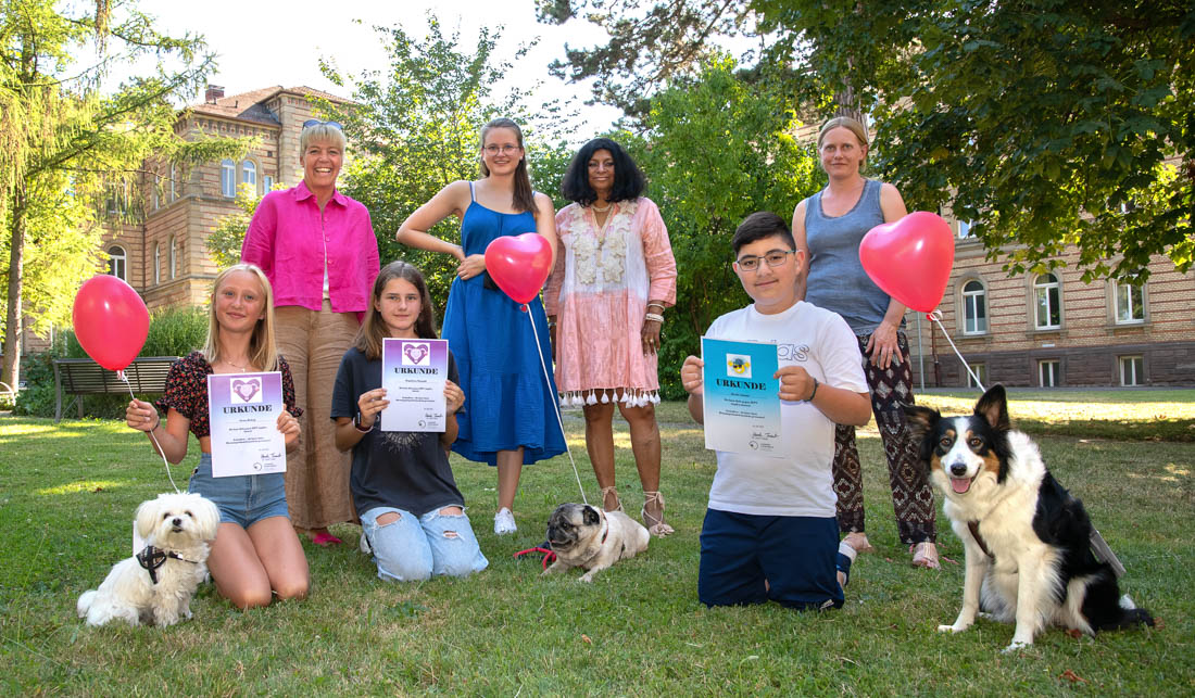Eine Gruppe von sieben Personen steht in einem Garten. Einige halten Luftballons und Zertifikate. Vorne sitzen zwei Hunde. Die Umgebung wirkt freundlich und festlich, mit einem Gebäude im Hintergrund, das zur Szene beiträgt.