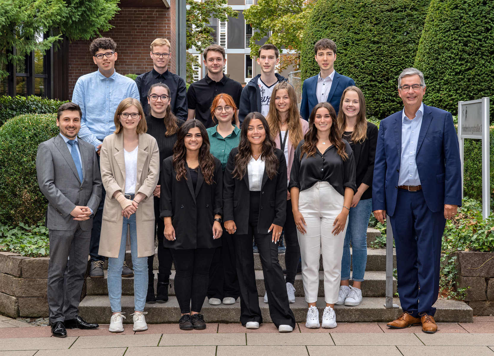 A group of 15 individuals stands on steps outside a building, arranged in three rows. They are dressed in professional attire, with a mix of suits and casual clothing. The setting features greenery in the background, creating a formal yet approachable atmosphere.