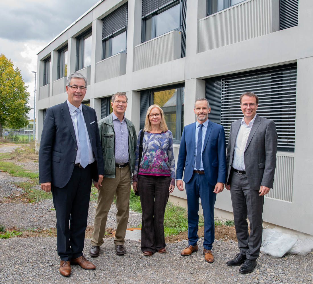 Fünf Personen stehen zusammen vor einem modernen Gebäude. Der Mann links trägt einen Anzug, gefolgt von einem Mann in legerer Kleidung, einer Frau mit blauer Bluse, einem weiteren Mann im Anzug und einem weiteren Mann in heller Kleidung. Die Gruppe lächelt.