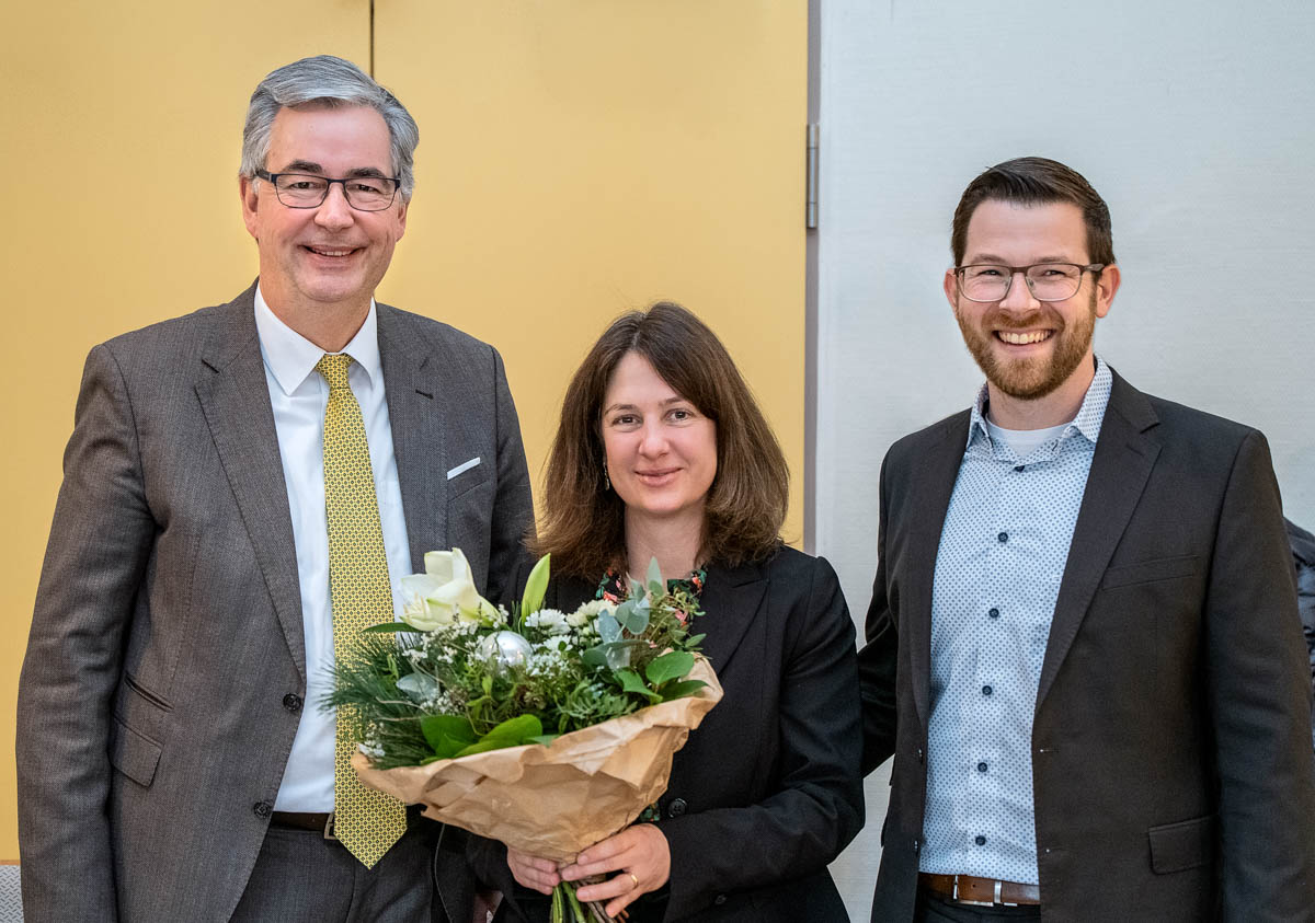 Trois personnes se tiennent ensemble, souriantes. Une femme au centre tient un bouquet de fleurs, tandis que deux hommes, un à sa gauche et l'autre à sa droite, l'entourent. La scène semble célébrer un moment spécial, possiblement une remise de prix ou une reconnaissance.
