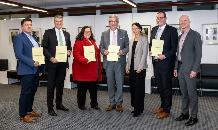 Eine Gruppe von sechs Personen steht in einem Raum und hält jeweils ein gelbes Dokument in den Händen. Sie zeigen sich freundlich und stehen in einer Reihe. Der Raum hat eine moderne Einrichtung mit Bildern an der Wand.
