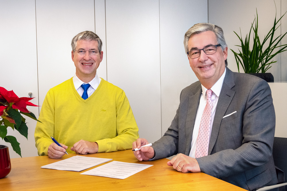 Deux hommes souriants sont assis à une table en bois. L'un porte un pull jaune et l'autre un costume gris avec une cravate rose. Ils tiennent des stylos et des documents signés, suggérant un accord ou une collaboration. Une plante verte et une fleur rouge sont visibles en arrière-plan.