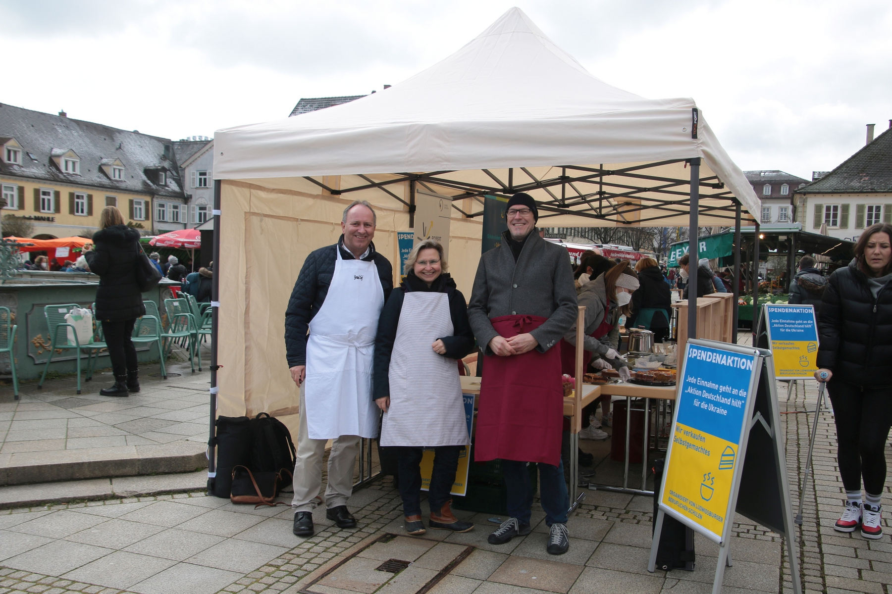 Drei Personen stehen lächelnd unter einem Marktstand mit einem weißen Zelt. Sie tragen Schürzen und präsentieren ihre Produkte. Im Hintergrund sind weitere Marktstände und eine belebte Platzatmosphäre sichtbar. Ein Informationsschild steht neben dem Stand.