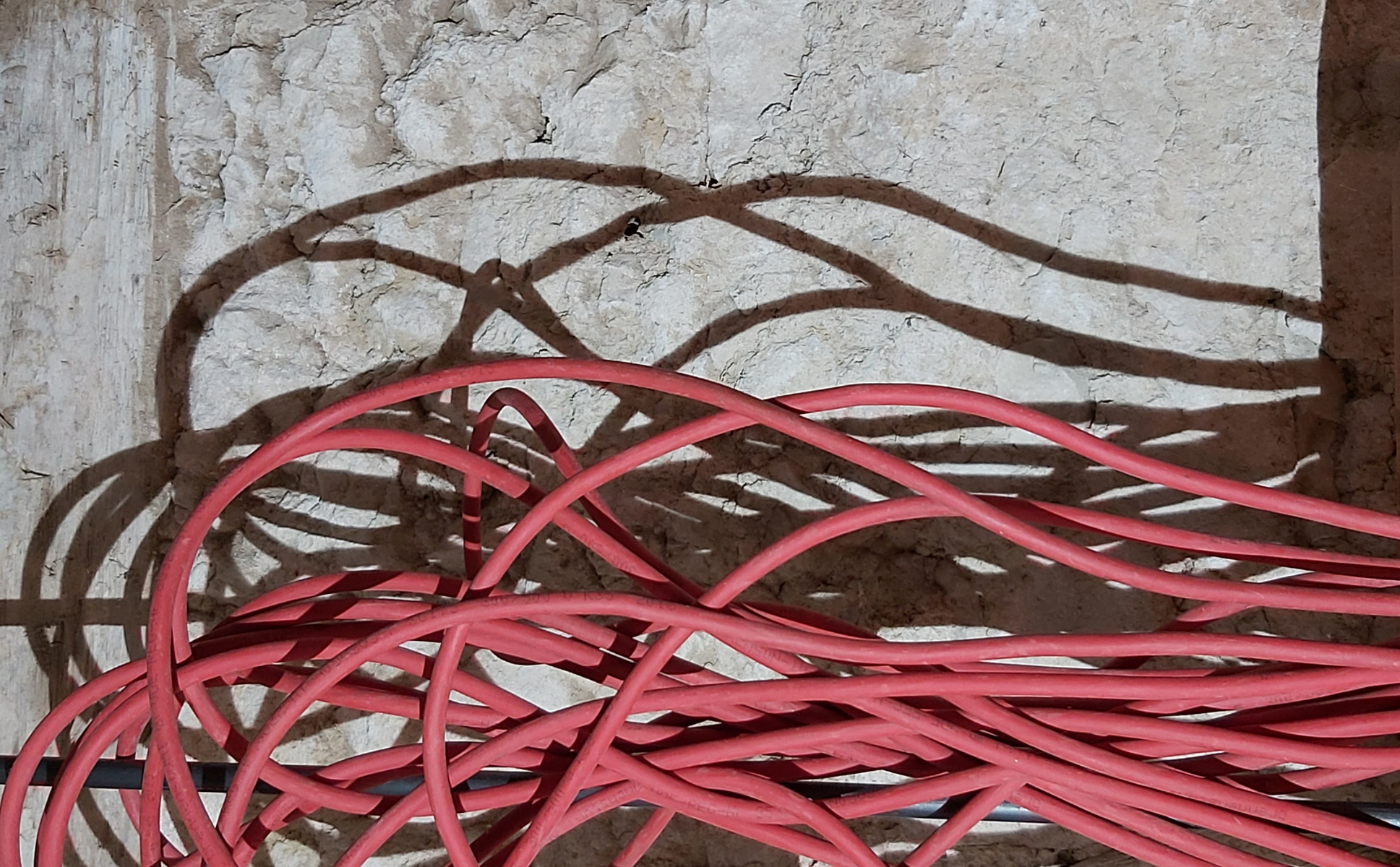 Auf dem Bild sind rote Kabel zu sehen, die wirr übereinanderliegen. Ihre Schatten bilden interessante Muster auf einer hellen, strukturierten Wand im Hintergrund. Die Kabel und deren Schatten verleihen dem Bild eine dynamische und künstlerische Note.