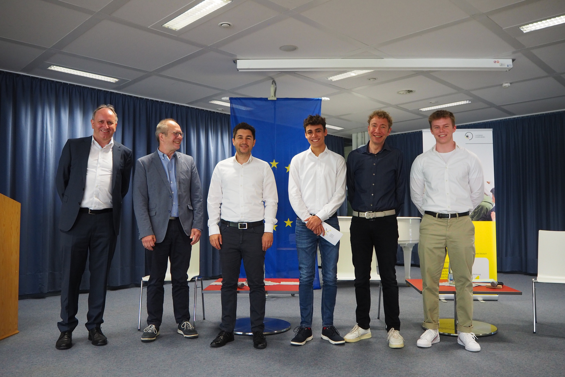 Sechs Männer stehen in einer Reihe in einem Konferenzraum. Sie tragen verschiedene Formate eleganter Kleidung und lächeln. Im Hintergrund ist eine EU-Flagge zu sehen. Der Raum wirkt professionell und festlich, passend zu einer besonderen Veranstaltung.