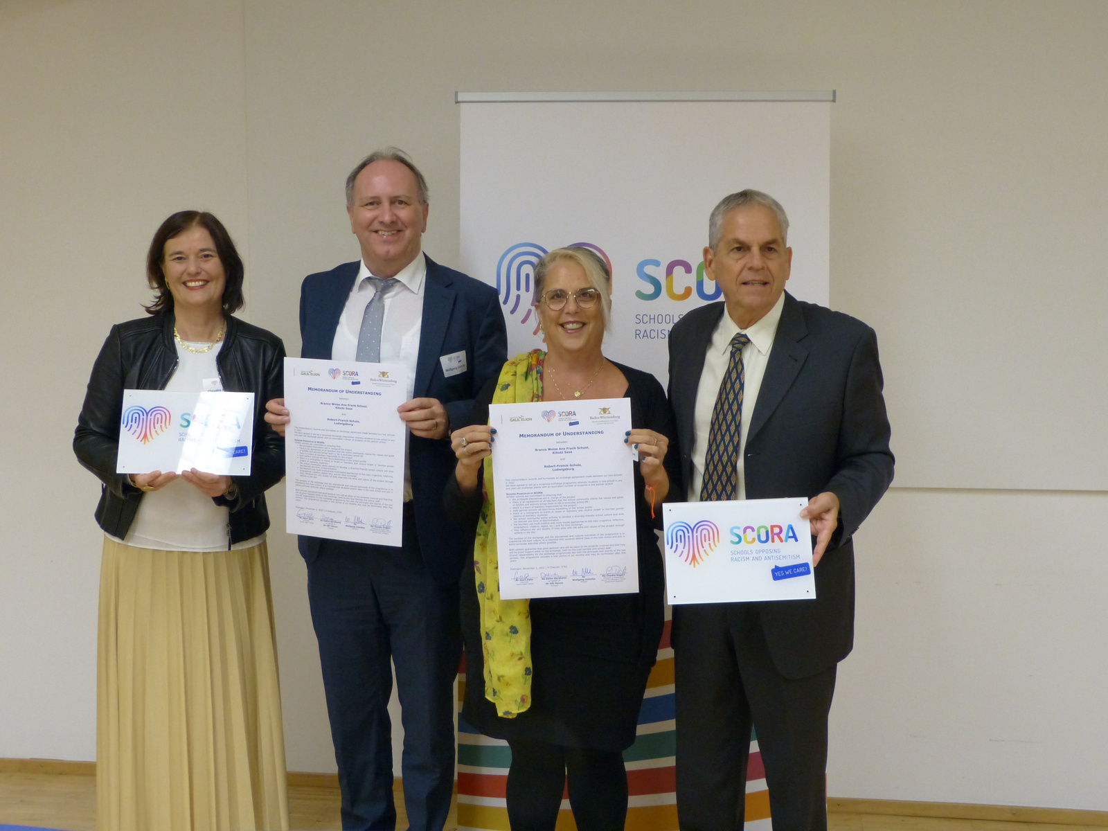 Quatre personnes se tiennent debout, tenant des certificats. Elles sourient, debout devant un panneau avec le logo de SCORA. L'image évoque une cérémonie officielle, axée sur la sensibilisation à la discrimination raciale.