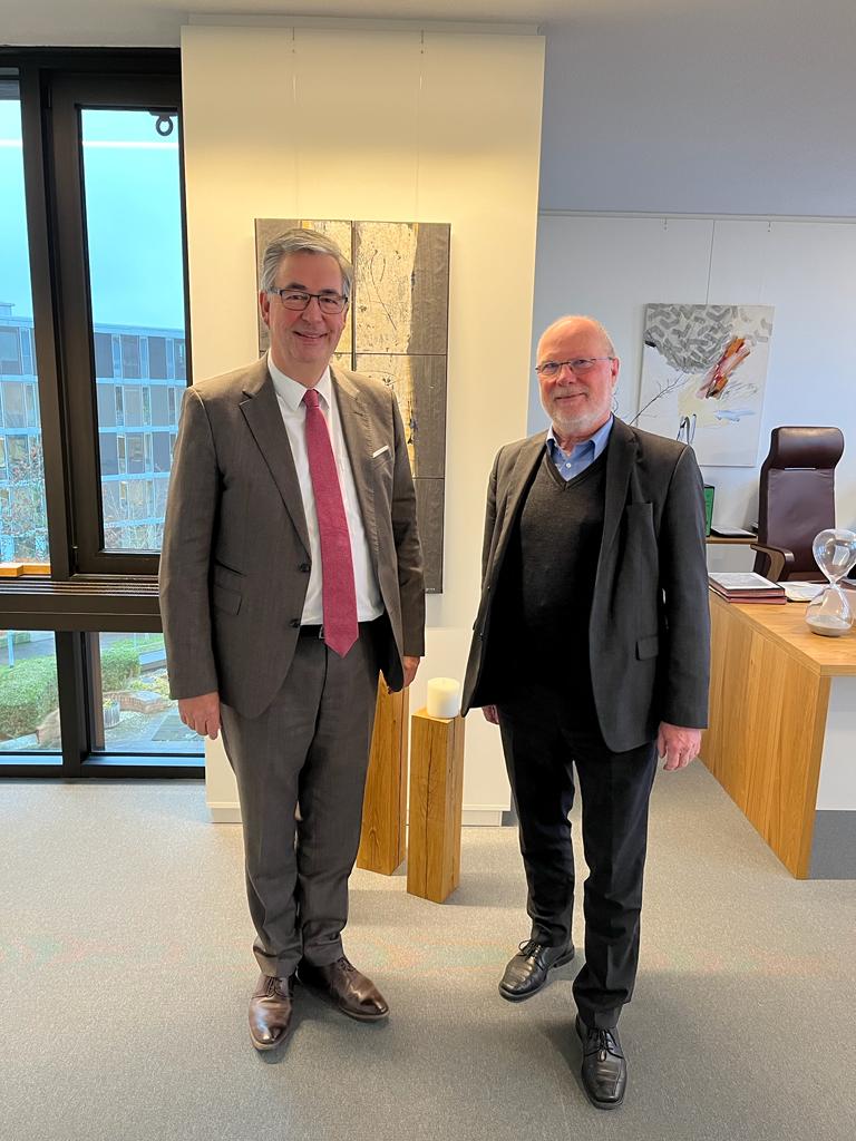 Due uomini in abiti formali si trovano in un ufficio. Uno indossa un completo grigio chiaro con cravatta, l'altro un completo scuro. Sullo sfondo si vede un tavolo da lavoro con elementi decorativi e una grande finestra che affaccia all'esterno.