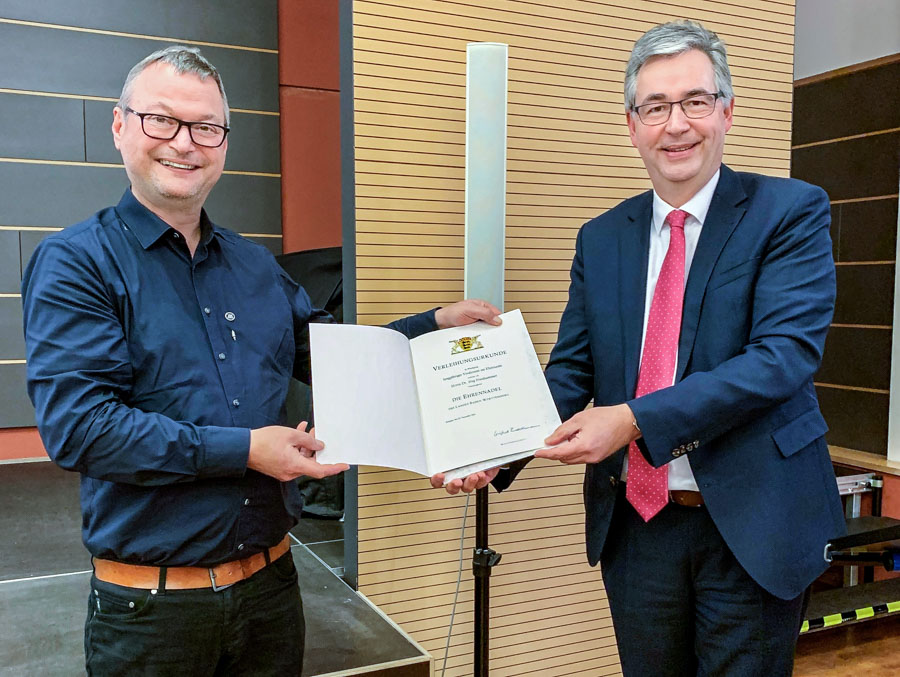 Zwei Männer stehen auf einer Bühne. Der linke Mann trägt ein schwarzes Hemd und lächelt, während er eine Urkunde in der Hand hält. Der rechte Mann trägt einen Anzug und präsentiert ebenfalls die Urkunde. Beide schauen lächelnd in Richtung Kamera.
